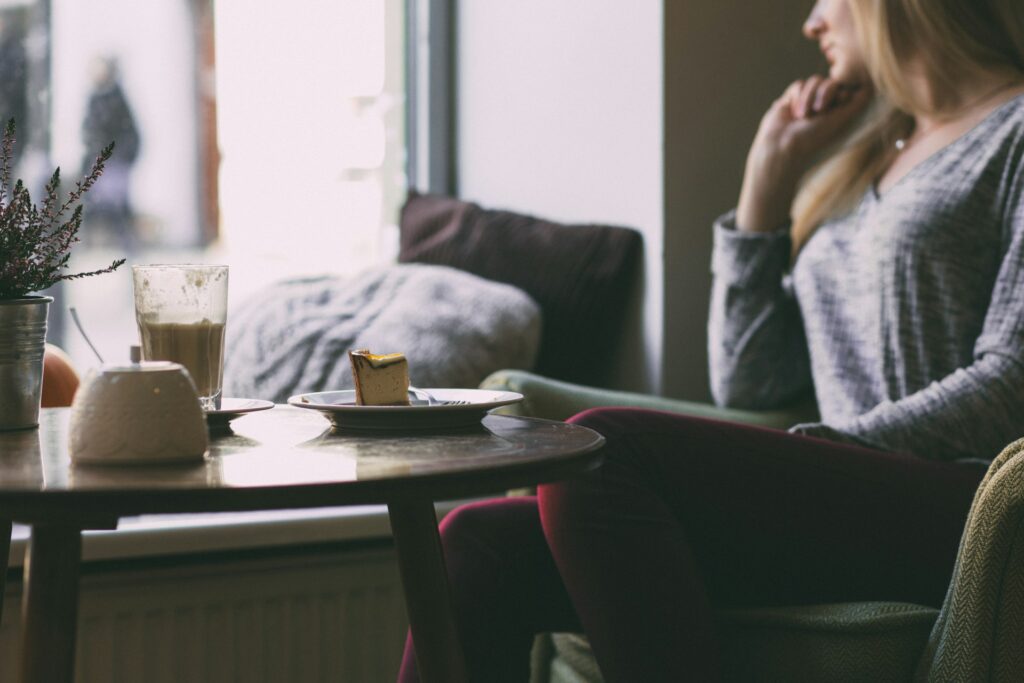 Frau sitzt im Café, denkt nach. Auf dem Tisch stehen ein Kaffee und ein Käsekuchen.